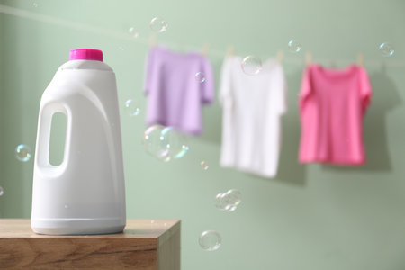 Bottle of fabric softener on wooden table and hanging clothes against pale dusty green background with soap bubbles, space for textの写真素材