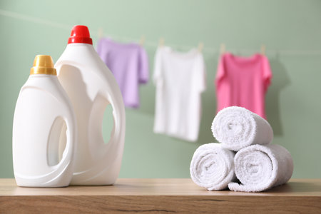 Bottles of fabric softener and towels on wooden table and hanging clothes against pale dusty green backgroundの写真素材