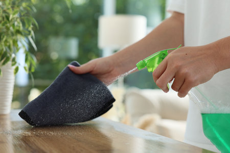 Woman spraying cleaning product while wiping wooden table with rag indoors, closeup
