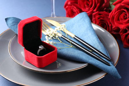 Beautiful table setting for romantic dinner, box with engagement ring and roses on blue background,...