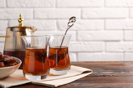 Tasty Turkish tea in glass cups, dates and teapot on wooden table against white brick wall, closeup. Space for textの写真素材