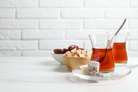 Tasty Turkish tea in glass cups, brown sugar and sweets on white marble table against brick wall, closeup. Space for textの写真素材