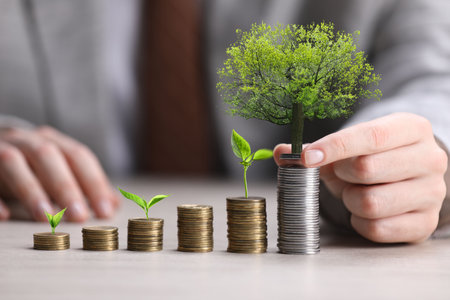 Wealth concept. Man stacking coins at table, closeup. Tree on bigger stack and seedlingsの写真素材