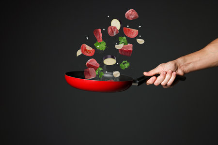 Chef tossing meat and vegetables pieces in frying pan against black background, closeupの写真素材