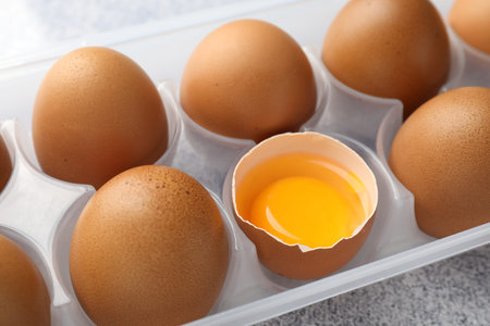 Whole Many raw chicken eggs and broken one in container on gray table, closeup