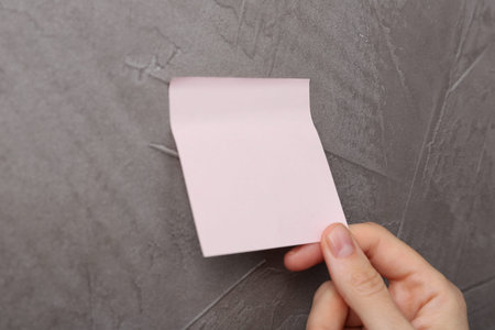 Woman putting blank adhesive note onto gray wall, closeup. Space for textの写真素材
