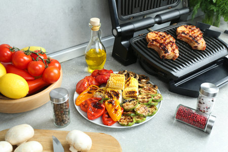 Electric grill with delicious steaks and vegetables on light table in kitchenの写真素材