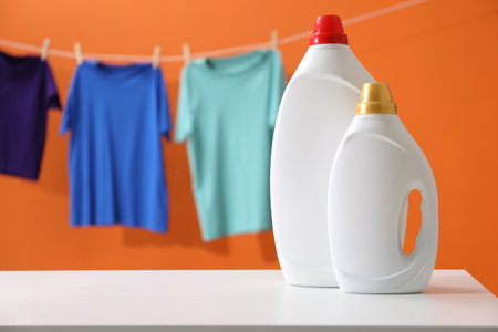 Bottles of fabric softener on white table and hanging clothes against orange background, space for textの写真素材