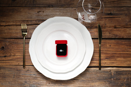 Beautiful table setting for romantic dinner and box with engagement ring on wooden background, flat...