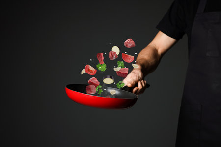 Chef tossing meat and vegetables pieces in frying pan against black background, closeupの写真素材