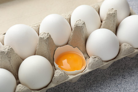 Many whole raw chicken eggs and broken one in carton on gray table, closeup