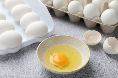 whole Many raw eggs and broken one in bowl on gray table, closeup