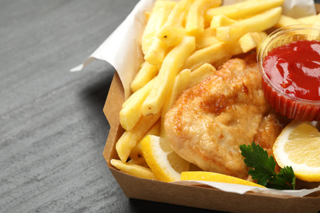 British Traditional Fish and chips with sauce, lemon and parsley in paper box on dark textured...