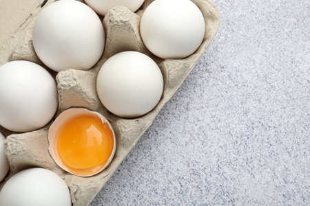 Many whole raw chicken eggs and broken one in carton on gray table, top view. Space for text