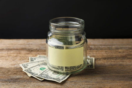 Jar with blank note and money on wooden table against black background