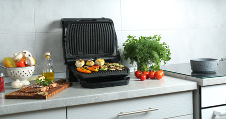 Electric grill with delicious vegetables and steaks on light table in kitchenの写真素材