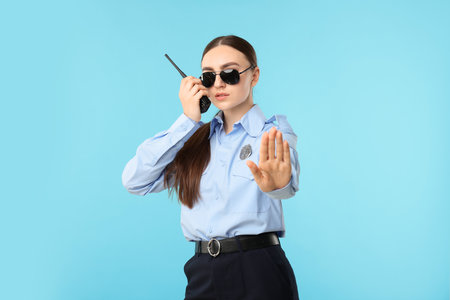 Young policewoman in uniform with walkie talkie showing stop gesture on light blue backgroundの写真素材