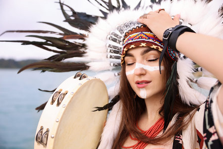 Beautiful shaman woman in native American Indian headdress with face painting and tambourine outdoors, closeupの写真素材