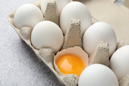 Many whole raw chicken eggs and broken one in carton on gray table, closeup