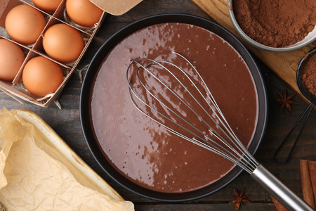 Liquid chocolate dough in bowl, ingredients on wooden table, flat lay