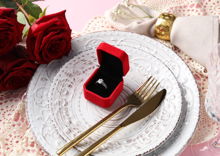 Beautiful table setting for romantic dinner, box with engagement ring and roses on pink background,...