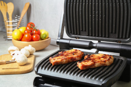 Electric grill with delicious steaks and vegetables on light table in kitchen, closeupの写真素材