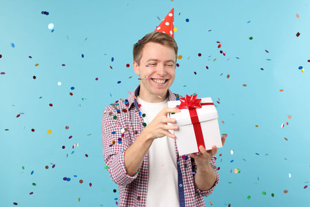 Happy young man with party hat and gift box among falling confetti on light blue backgroundの写真素材
