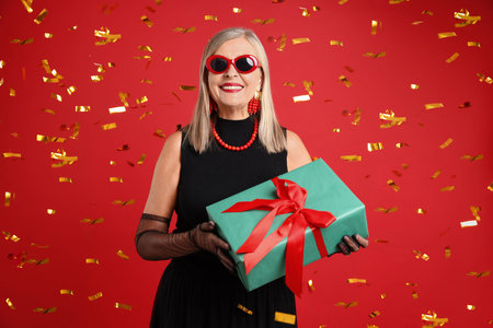 Happy senior woman with gift and flying confetti against red backgroundの写真素材