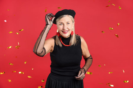 Happy senior woman in elegant outfit and flying confetti against red backgroundの写真素材