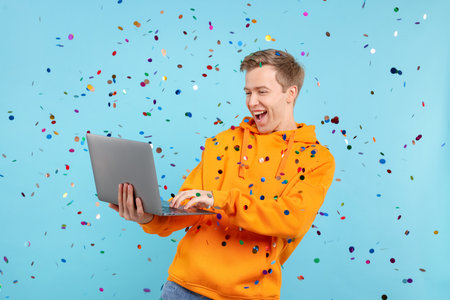 Excited young man with laptop among falling confetti on light blue backgroundの写真素材