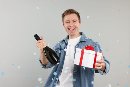 Happy young man with gift box and bottle of sparkling wine among falling confetti on light gray backgroundの写真素材