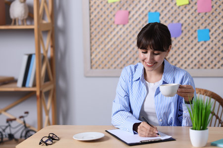 Coffee break. Smiling woman writing with cup at wooden table indoors, space for textの写真素材