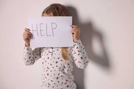 Sad little girl holding sheet of paper with word Help near white wall indoors, space for textの写真素材