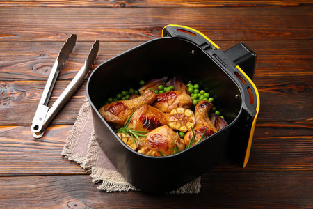 Air fryer basket with grilled chicken drumsticks, spices, green peas and tongs on wooden table, closeupの写真素材
