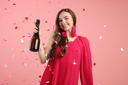 Happy young woman with bottle of sparkling wine among falling confetti on pink backgroundの写真素材