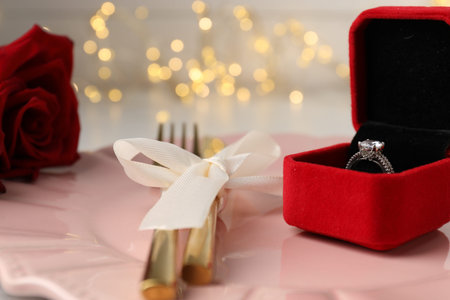 Romantic place setting. Box with engagement ring, plate, cutlery and rose against blurred lights,...