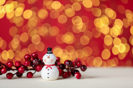 Cute ceramic snowman and decorative branch with berries on white table against red background with blurred lights, closeup. Space for textの写真素材