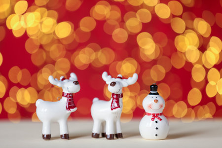 Cute decorative snowman and reindeers on white table against red background with blurred lights, closeup. Space for textの写真素材