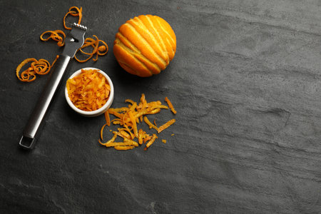 Orange zest, fresh fruit and zester tool on black table, flat lay.