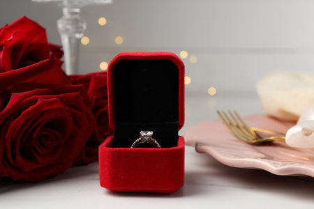 Romantic place setting. Box with engagement ring, plate, cutlery and roses on white table, closeup
