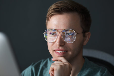 Smiling man wearing glasses with reflection working with computer on gray background