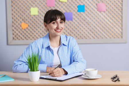 Coffee break. Smiling woman with clipboard, pen and cup at wooden table indoorsの写真素材