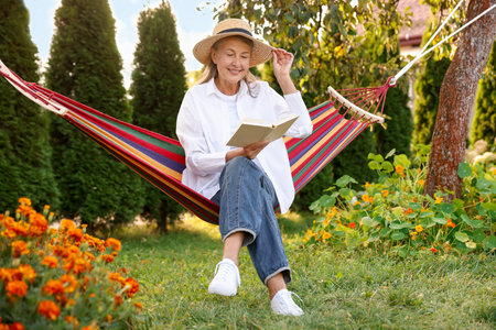 Senior woman with straw hat and book resting in hammock outdoorsの写真素材