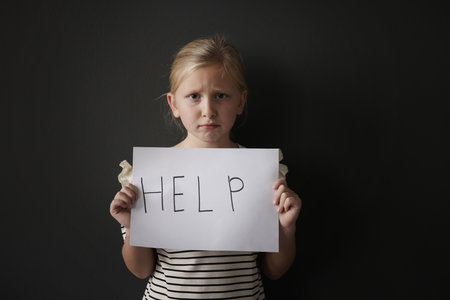 Sad little girl holding sheet of paper with word Help on black backgroundの写真素材