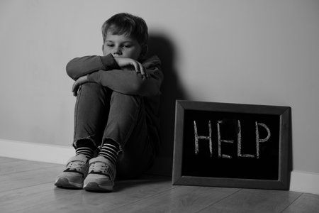 Upset little boy and small chalkboard with the word Help near wall indoors, black and white effectの写真素材