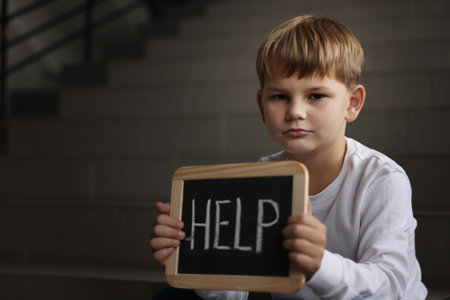 Upset little boy holding small chalkboard with word Help on stairs indoors, space for textの写真素材