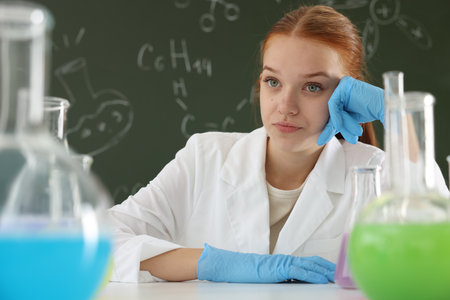 Back to school. Girl at desk with flasks of liquids against chalkboard with chemical formulas indoorsの写真素材