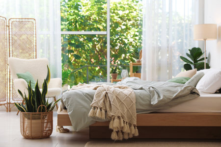 Cozy large bed with lines, houseplants and lamp near window in bedroom. Interior designの写真素材