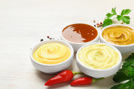 Different tasty sauces in bowls, parsley, peppercorns and chili peppers on white wooden table, closeup. Space for textの写真素材