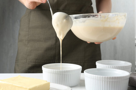 Woman pouring batter (liquid dough) into baking dishes at table indoors, closeup